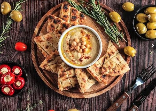 Hummus with grilled pitta wedges, kalamata olives and stuffed peppers on a wooden board